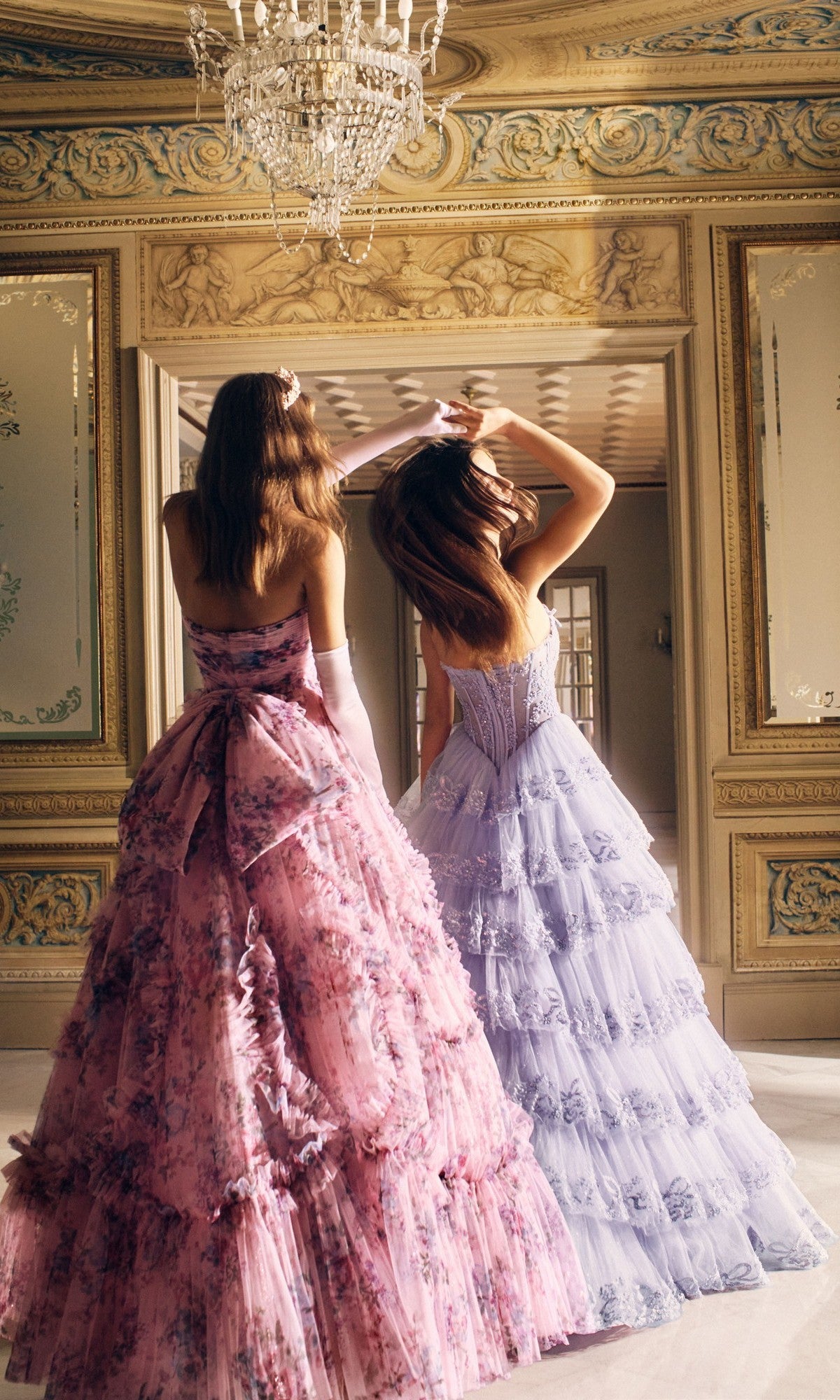 Two women stand in an elegant room with gilded walls and a chandelier, both wearing the Alyce 62191 long prom dress—one in a corset bodice with pink floral details, the other in a lavender tulle A-line skirt—facing away from the camera.